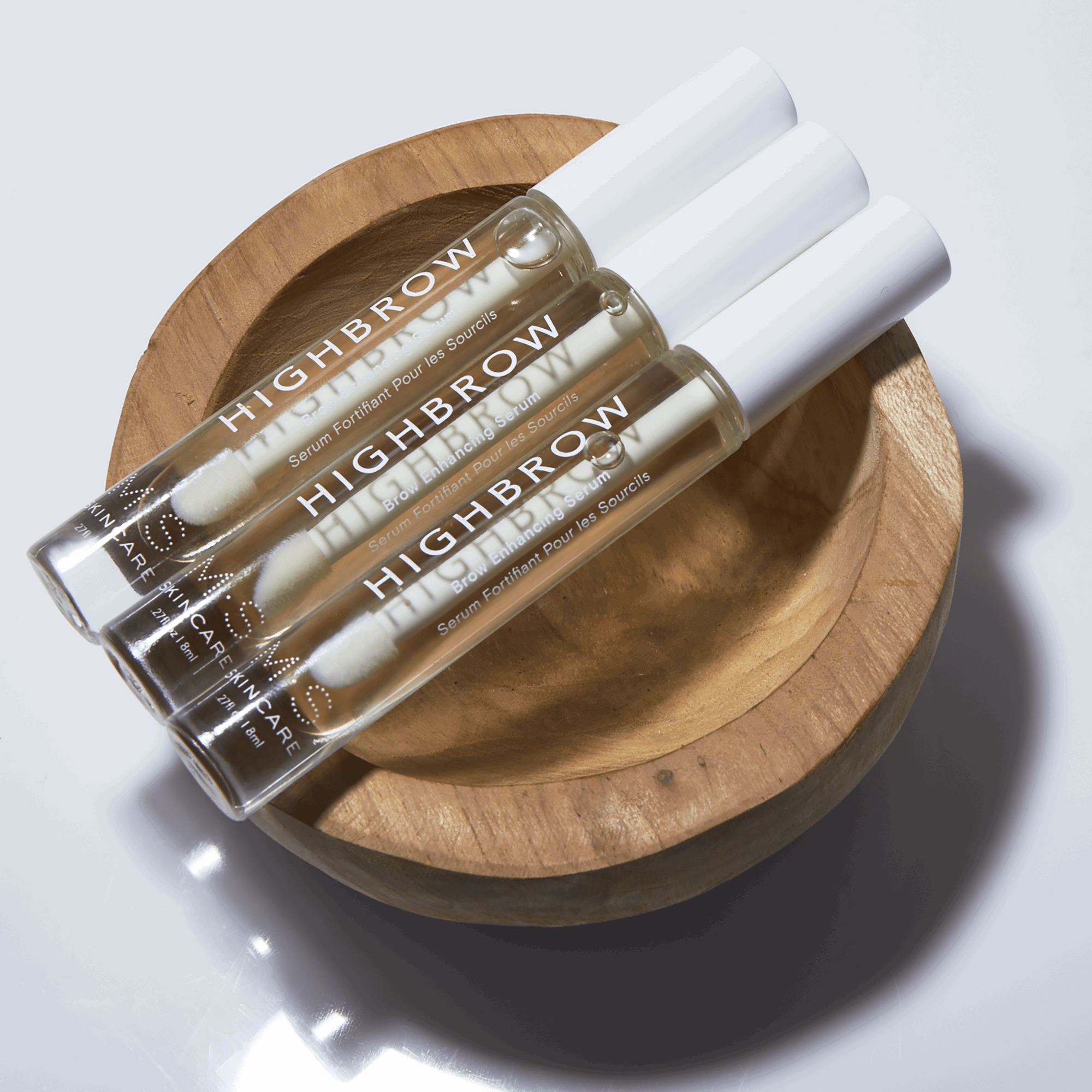 M.S Skincare HIGHBROW Brow Enhancing Serum in a glass container with white wand and cap- 3 units on top of a wooden bowl.